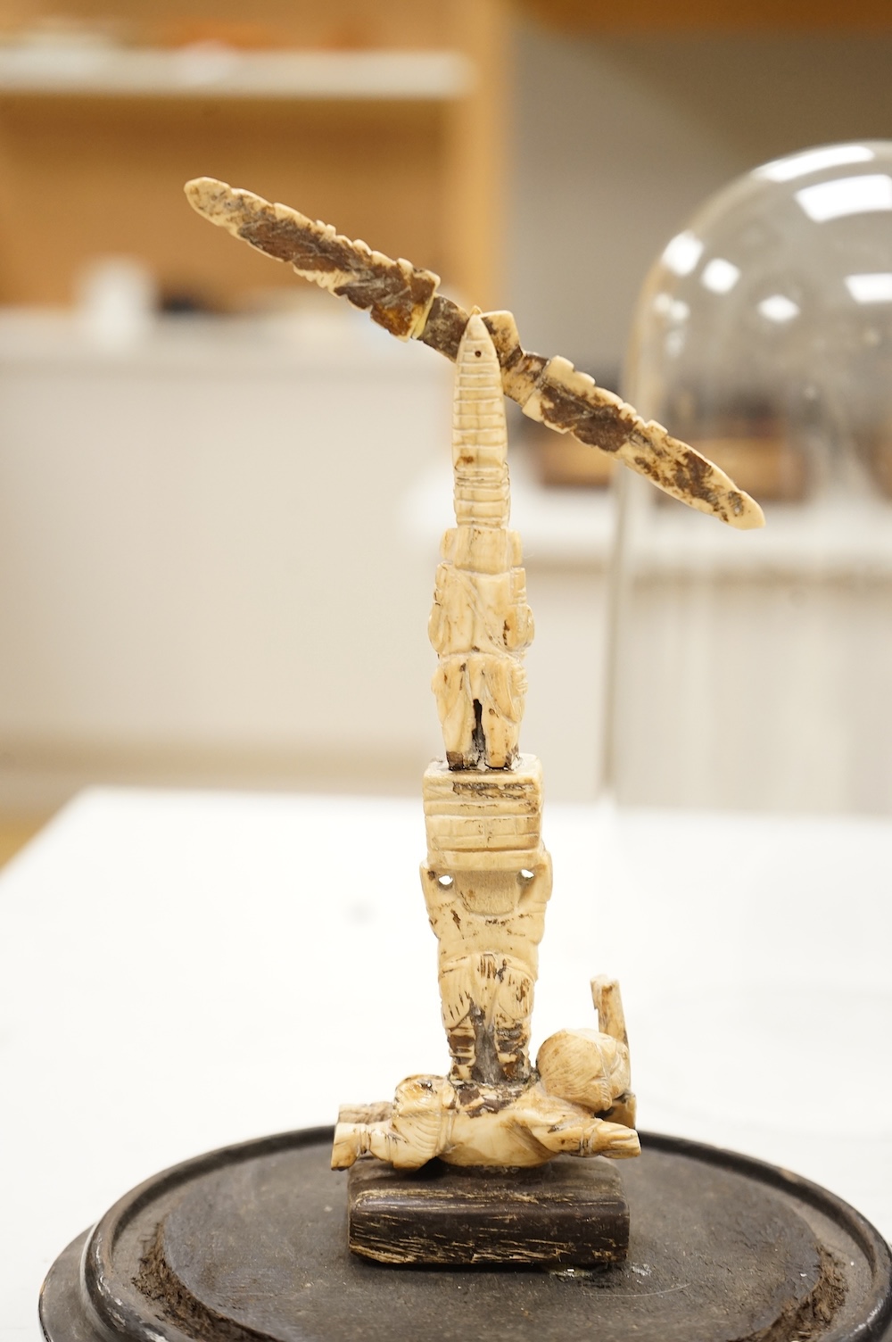 A 19th century Indian bone sculptural devotional object, carved as a totem with Ganesh and other deities, pivoting beam, 18cm high, glass dome with ebonised base